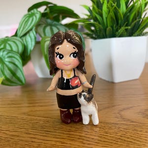 May include: A handcrafted figurine of a person with brown hair, wearing a black top and skirt, and maroon boots. A small, white and brown cat stands beside the figurine. The figurine is on a wooden surface, with green plants in the background.