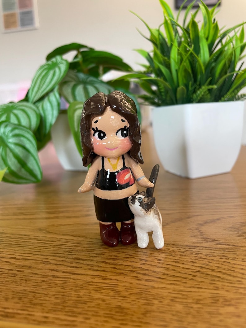 May include: A handcrafted figurine of a person with brown hair, wearing a black top and skirt, and maroon boots. A small, white and brown cat stands beside the figurine. The figurine is on a wooden surface, with green plants in the background.