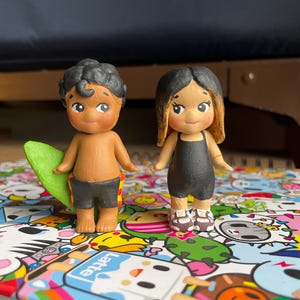 May include: Two small, hand-painted figurines of a boy and a girl. The boy is holding a green surfboard and wearing black shorts with a yellow and red pattern. The girl is wearing a black swimsuit and sandals. Both have tan skin and black hair.