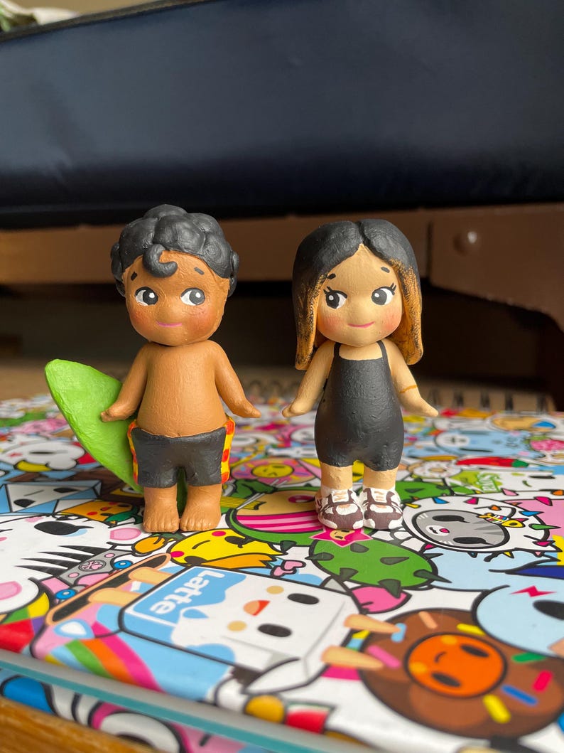 May include: Two small, hand-painted figurines of a boy and a girl. The boy is holding a green surfboard and wearing black shorts with a yellow and red pattern. The girl is wearing a black swimsuit and sandals. Both have tan skin and black hair.