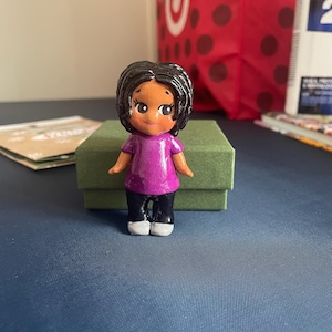 May include: A hand-painted figurine of a person with dark hair, wearing a purple shirt, black pants, and white shoes. The figurine is standing on a green box. The background includes a red shopping bag with white polka dots.