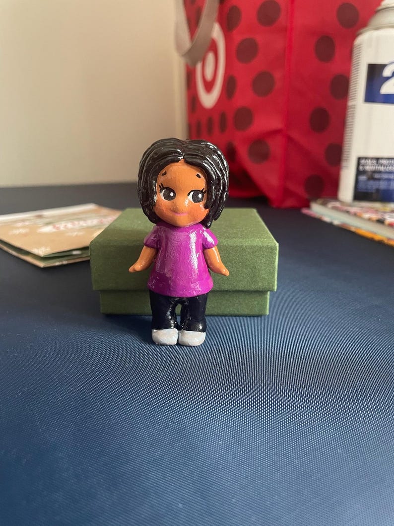 May include: A hand-painted figurine of a person with dark hair, wearing a purple shirt, black pants, and white shoes. The figurine is standing on a green box. The background includes a red shopping bag with white polka dots.