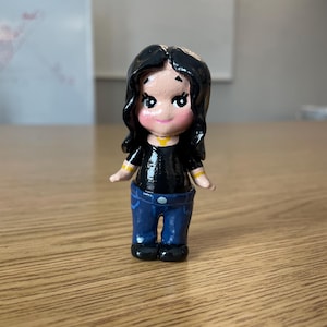 May include: A small, hand-painted figurine of a person with black hair, wearing a black top, blue jeans, and black shoes. The figurine has a yellow collar and yellow stripes on the sleeves. The figure is standing on a wooden surface.