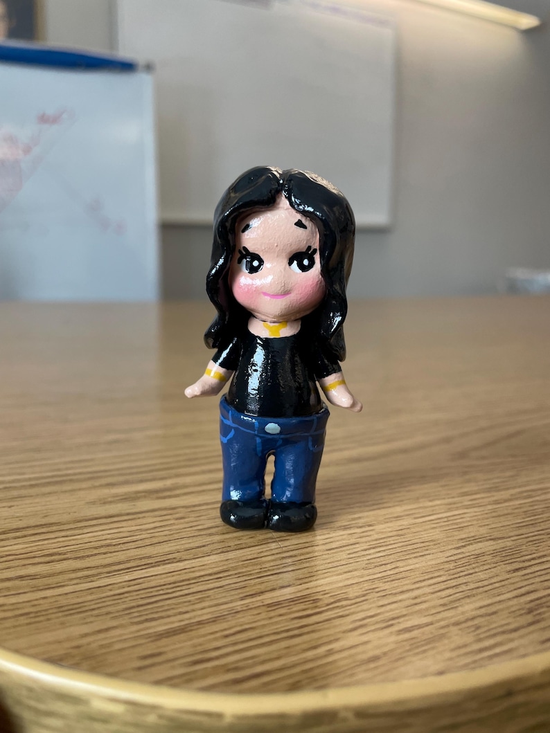 May include: A small, hand-painted figurine of a person with black hair, wearing a black top, blue jeans, and black shoes. The figurine has a yellow collar and yellow stripes on the sleeves. The figure is standing on a wooden surface.