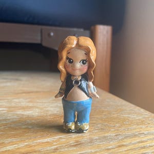 May include: A handcrafted figurine of a person with red hair, wearing a black top, blue jeans, and gold shoes. The figurine has a detailed face with freckles and tattoos on the arms. The figure is standing on a wooden surface.