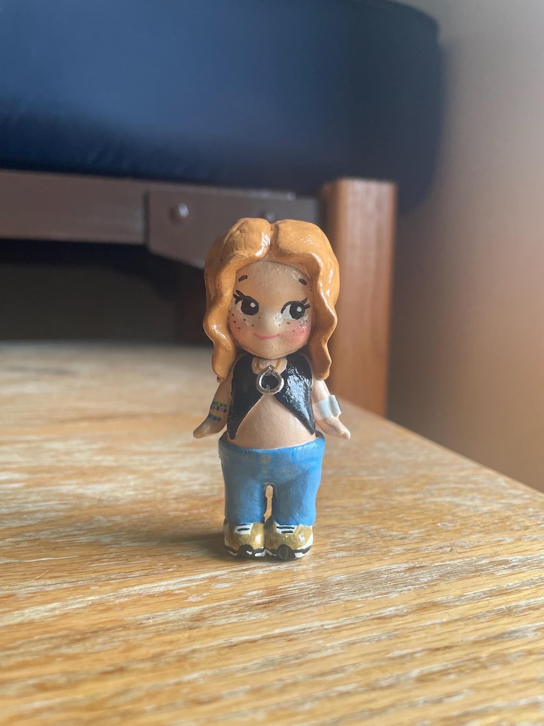 May include: A handcrafted figurine of a person with red hair, wearing a black top, blue jeans, and gold shoes. The figurine has a detailed face with freckles and tattoos on the arms. The figure is standing on a wooden surface.