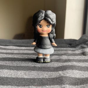 May include: A small, handcrafted figurine of a girl with black hair, wearing a black top and a gray skirt. The doll has rosy cheeks and is wearing black shoes with yellow accents. The figurine is standing on a gray and white striped surface.