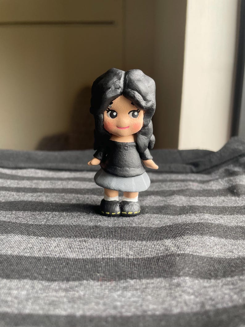 May include: A small, handcrafted figurine of a girl with black hair, wearing a black top and a gray skirt. The doll has rosy cheeks and is wearing black shoes with yellow accents. The figurine is standing on a gray and white striped surface.