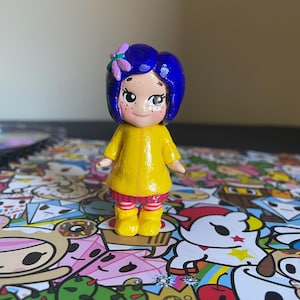 May include: A handcrafted figurine of a character with blue hair, a yellow dress, and yellow boots. The figurine has a flower in its hair and freckles on its face. The figure is standing on a colorful patterned surface.