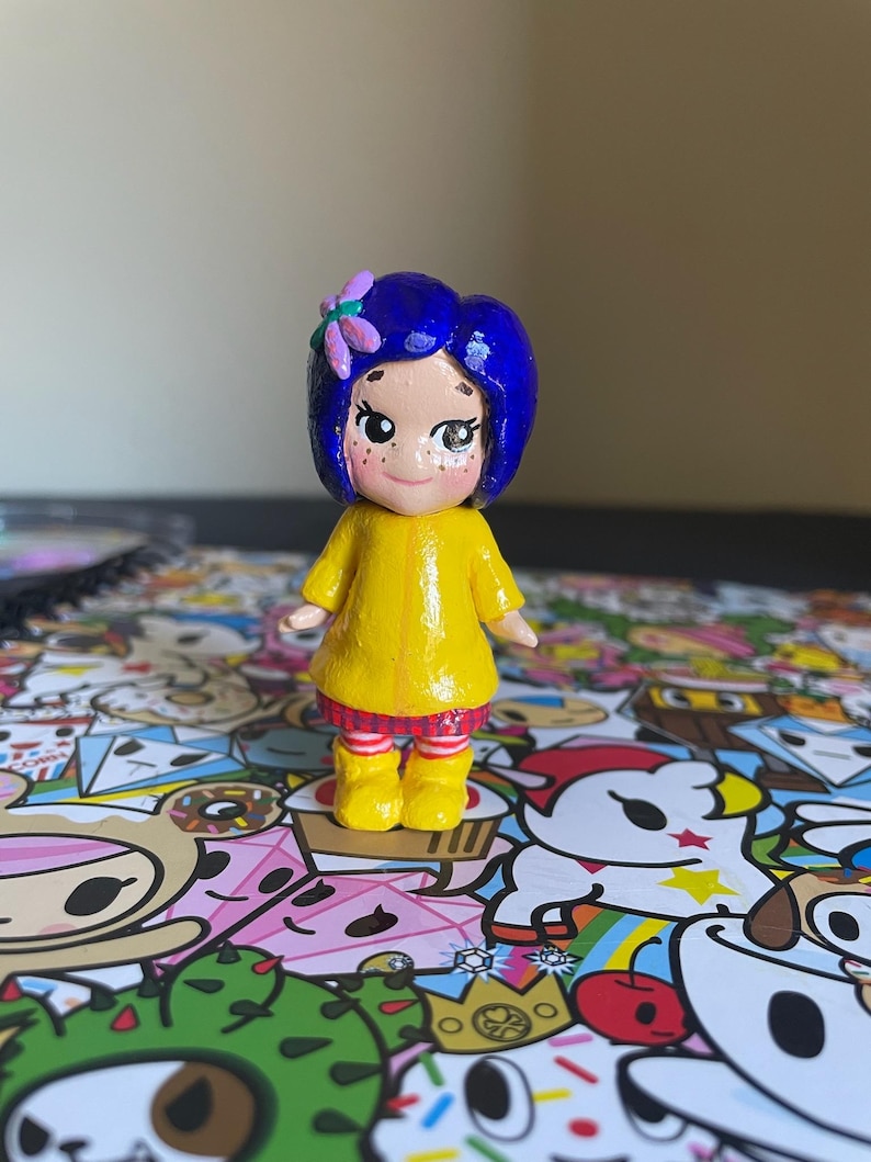 May include: A handcrafted figurine of a character with blue hair, a yellow dress, and yellow boots. The figurine has a flower in its hair and freckles on its face. The figure is standing on a colorful patterned surface.