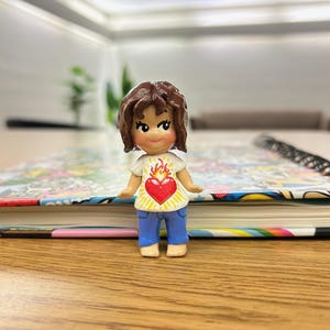 May include: A small figurine of a person with brown hair, wearing a white shirt with a red heart and flame design, and blue pants. The figurine is standing on a wooden surface, with a colorful book in the background.