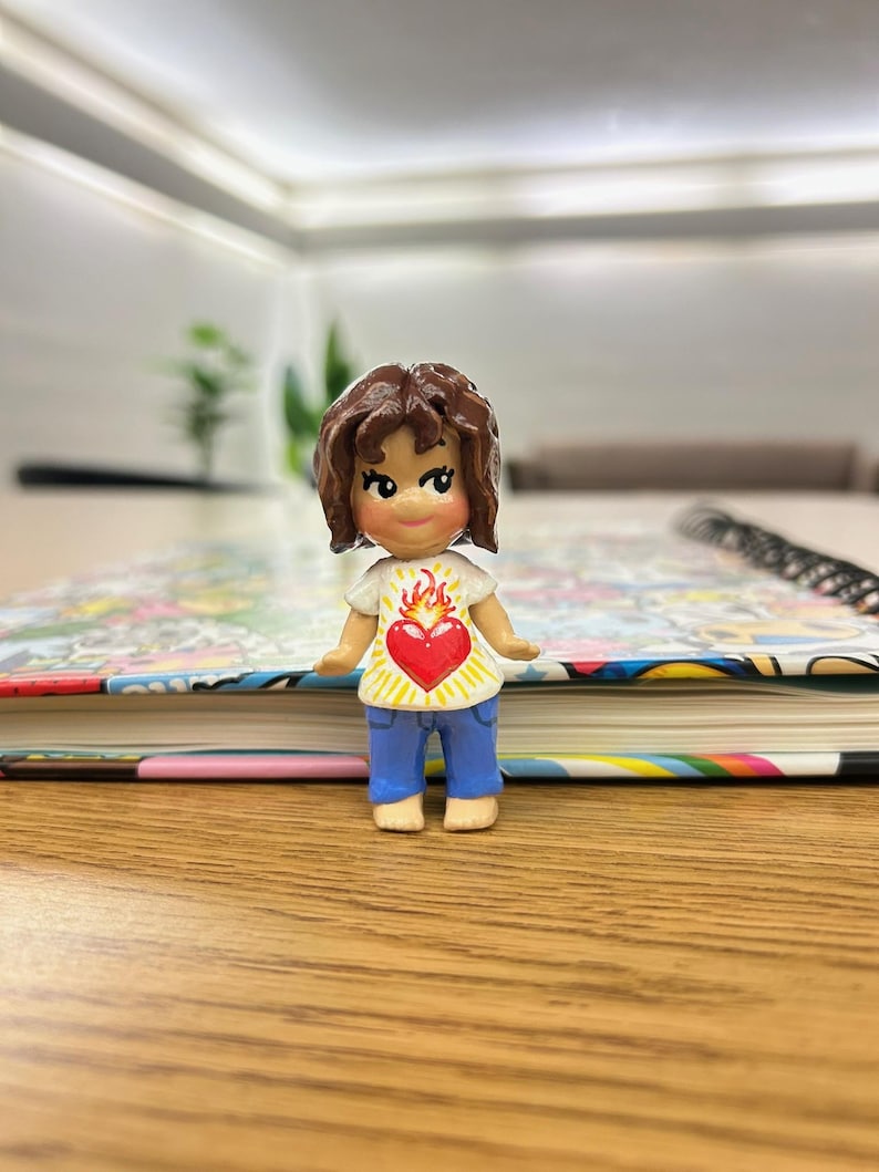 May include: A small figurine of a person with brown hair, wearing a white shirt with a red heart and flame design, and blue pants. The figurine is standing on a wooden surface, with a colorful book in the background.