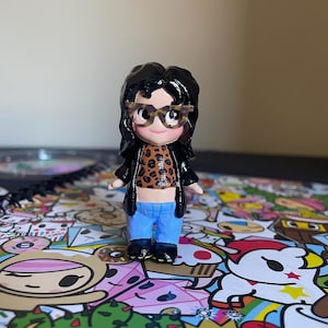 May include: A small figurine of a person wearing glasses, a leopard print top, a black jacket, and blue jeans. The figurine has black hair and is standing on a colorful patterned surface.