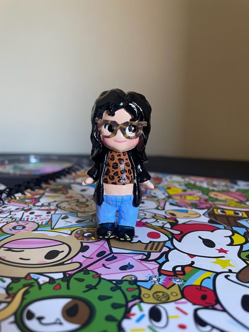 May include: A small figurine of a person wearing glasses, a leopard print top, a black jacket, and blue jeans. The figurine has black hair and is standing on a colorful patterned surface.