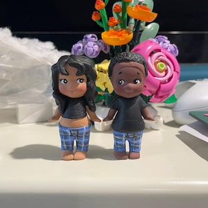 May include: Two small, hand-painted figurines of a couple. The figures are wearing black tops and blue plaid pants. The woman has black curly hair, and the man has short black hair. They are standing on a white surface.