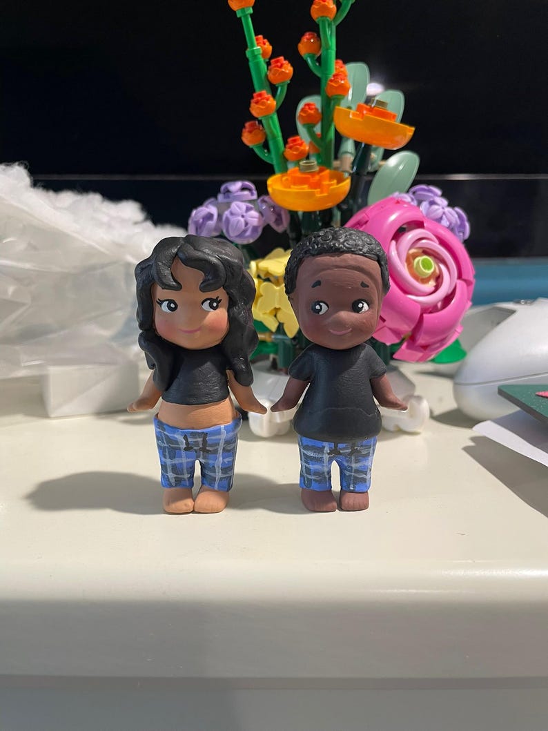 May include: Two small, hand-painted figurines of a couple. The figures are wearing black tops and blue plaid pants. The woman has black curly hair, and the man has short black hair. They are standing on a white surface.