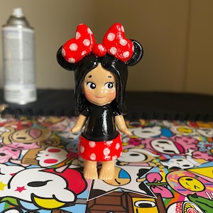 May include: A hand-painted figurine of a character with black hair, a red bow with white polka dots, a black top, and a red skirt with white polka dots. The figurine is standing on a colorful surface.