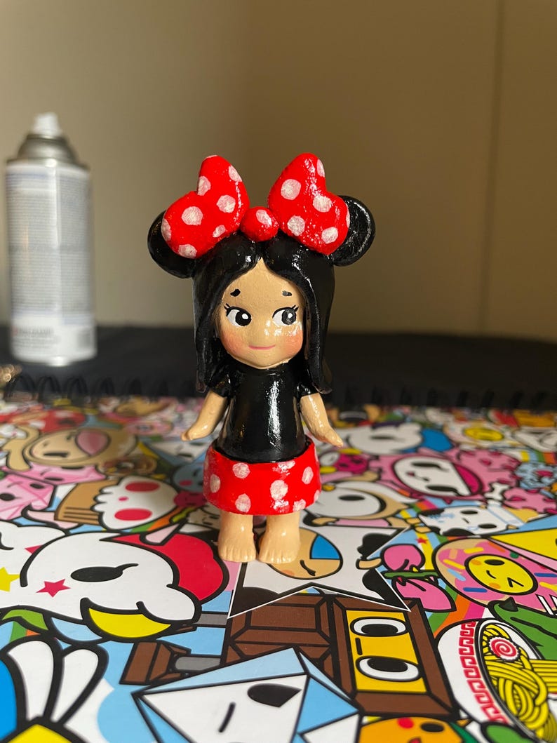 May include: A hand-painted figurine of a character with black hair, a red bow with white polka dots, a black top, and a red skirt with white polka dots. The figurine is standing on a colorful surface.
