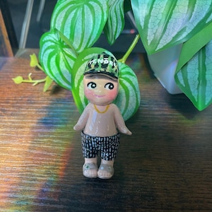 May include: A small, hand-painted ceramic figurine of a person wearing a cap with the word "FENDI" on it, a tan shirt, and black and white patterned shorts. The figurine has a gold necklace and gray sandals. The background includes green plants.