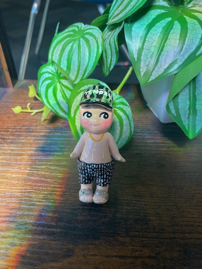 May include: A small, hand-painted ceramic figurine of a person wearing a cap with the word "FENDI" on it, a tan shirt, and black and white patterned shorts. The figurine has a gold necklace and gray sandals. The background includes green plants.