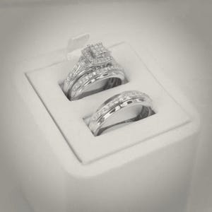 Puede incluir: Un conjunto de anillos plateados presentados en una caja blanca. El anillo de compromiso presenta un grupo cuadrado de pequeñas piedras, con piedras adicionales a lo largo de la banda. La alianza es una banda simple con una fila de piedras.
