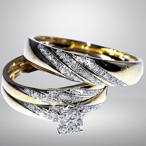 Op de afbeelding: Een set van twee goudkleurige ringen. Eén ring heeft een vierkante cluster van kleine diamanten, de andere heeft een diagonaal patroon van diamanten. De ringen zijn waarschijnlijk voor een bruiloft of verloving.