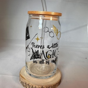 Könnte beinhalten: Ein klares Glas mit einem Bambusdeckel und einem Glasstrohhalm. Der Becher hat schwarze und goldene Grafiken und den Text "Theres a little MAGIC in all of us." Er steht auf einem Holzuntersetzer.