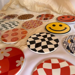 Könnte beinhalten: Eine Sammlung dekorativer Keramikuntersetzer mit verschiedenen Mustern und Designs. Die Untersetzer weisen eine Reihe von Farben auf, darunter Rot, Gelb, Schwarz und Weiß. Zu den Designs gehören Smiley-Gesichter, Schachbrettmuster, Blumenmotive und geometrische Formen.