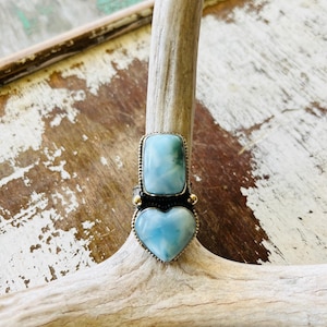 May include: A silver ring with two light blue gemstones. The top stone is rectangular, and the bottom stone is heart-shaped. The ring is set on a light-colored wooden surface.