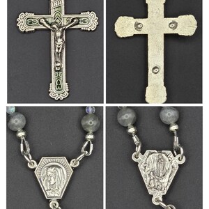 May include: A silver rosary with a detailed crucifix depicting Jesus. The rosary features grey gemstone beads and a pendant with a religious image. The cross has green enamel detailing.