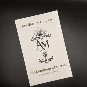 Pode incluir: Um folheto branco intitulado "Meditation Guide to the Luminous Mysteries" com ilustrações e texto decorativos em preto. O folheto apresenta as iniciais "AM" e um símbolo de cálice. O texto "traditionally prayed on Thursdays" também é visível.