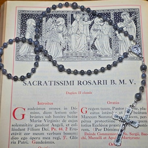 May include: A grey beaded rosary with a silver crucifix and a small religious pendant lies on an open book. The book has illustrations and Latin text, including "SACRATISSIMI ROSARII B. M. V."