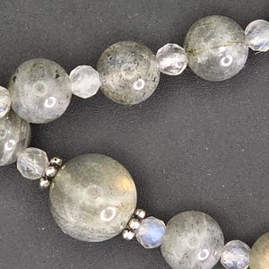 May include: A close-up shot of a necklace with large, round, grey-green labradorite beads and smaller, clear, faceted beads. The necklace is set against a dark background, showcasing the natural variations in the stones.