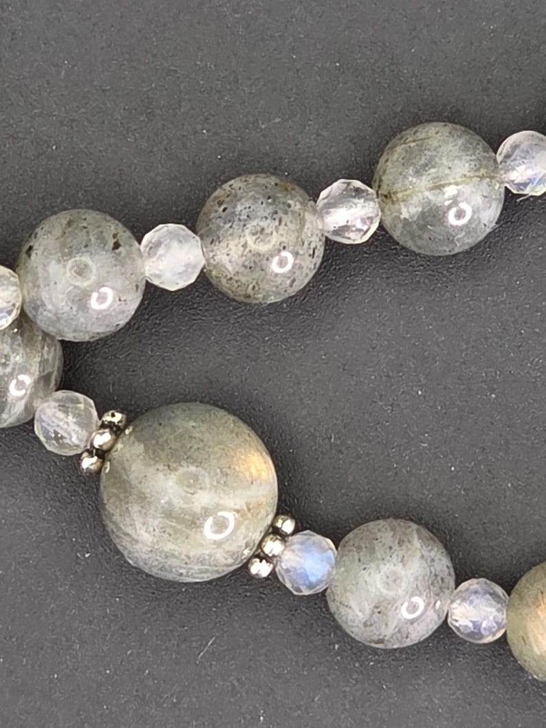 May include: A close-up shot of a necklace with large, round, grey-green labradorite beads and smaller, clear, faceted beads. The necklace is set against a dark background, showcasing the natural variations in the stones.