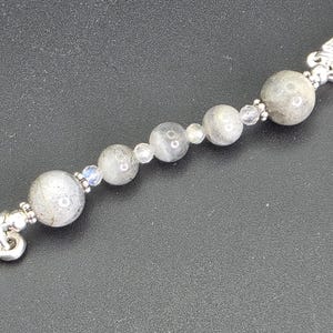 May include: A silver bracelet with a series of grey, spherical beads and smaller, clear beads. The bracelet has decorative silver end pieces. The beads have a speckled appearance.