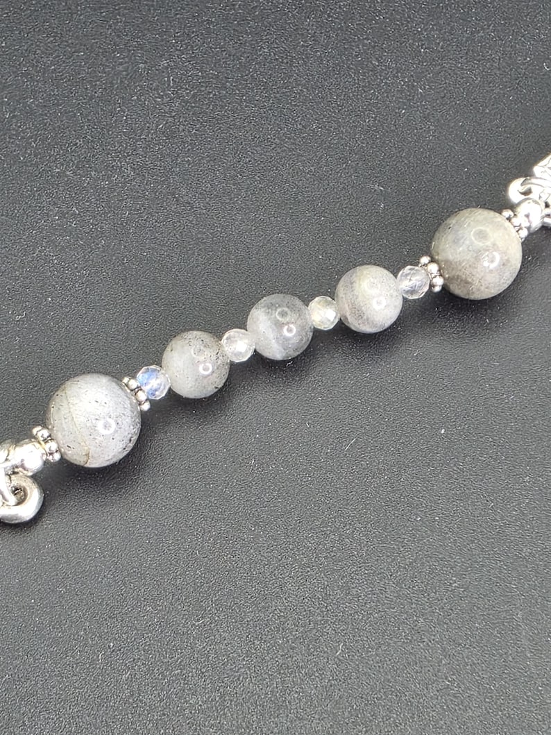 May include: A silver bracelet with a series of grey, spherical beads and smaller, clear beads. The bracelet has decorative silver end pieces. The beads have a speckled appearance.