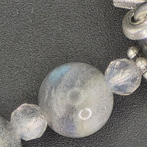 May include: Close-up of a bracelet featuring large, round, iridescent labradorite beads and faceted clear crystal beads. The bracelet also includes silver-toned metal accents and a decorative clasp.