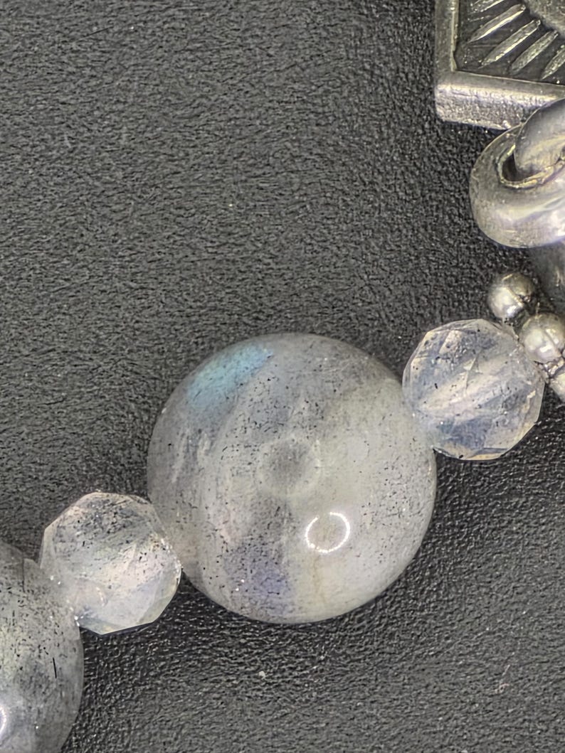 May include: Close-up of a bracelet featuring large, round, iridescent labradorite beads and faceted clear crystal beads. The bracelet also includes silver-toned metal accents and a decorative clasp.