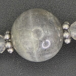 May include: Close-up of a beaded necklace. It features a large, round, grey bead with a speckled appearance. The bead is flanked by smaller, clear beads and silver-toned metal accents. The background is a dark, textured surface.