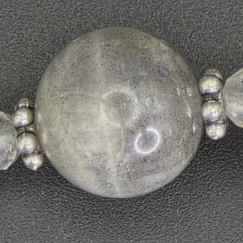 May include: Close-up of a beaded necklace. It features a large, round, grey bead with a speckled appearance. The bead is flanked by smaller, clear beads and silver-toned metal accents. The background is a dark, textured surface.