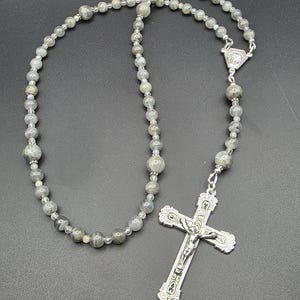 May include: A rosary featuring grey gemstone beads and a silver crucifix. The beads are of varying sizes and are strung together with a silver chain. The crucifix has a detailed depiction of Jesus Christ.