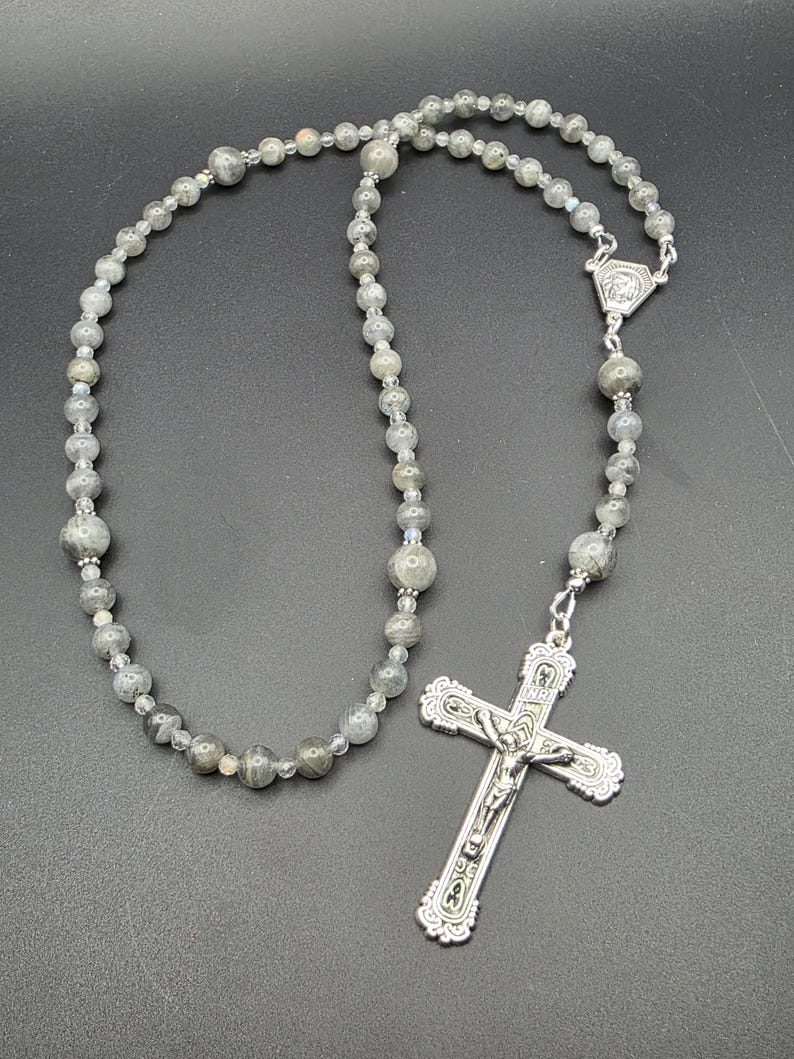 May include: A rosary featuring grey gemstone beads and a silver crucifix. The beads are of varying sizes and are strung together with a silver chain. The crucifix has a detailed depiction of Jesus Christ.