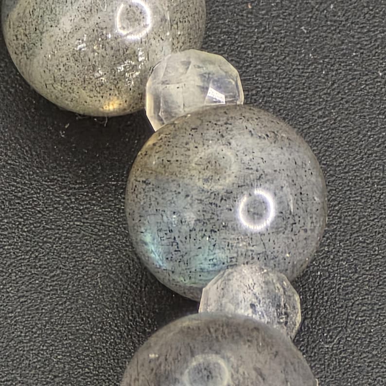May include: Close-up of a beaded necklace featuring large, round, grey-green labradorite beads interspersed with smaller, faceted, clear quartz beads. The labradorite beads have a speckled appearance and a subtle iridescence. The background is a dark, textured surface.