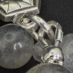 May include: Close-up of a silver bracelet with a decorative clasp and chain links. The bracelet features multiple round, textured beads. The metal has a polished finish, reflecting light against a dark background.