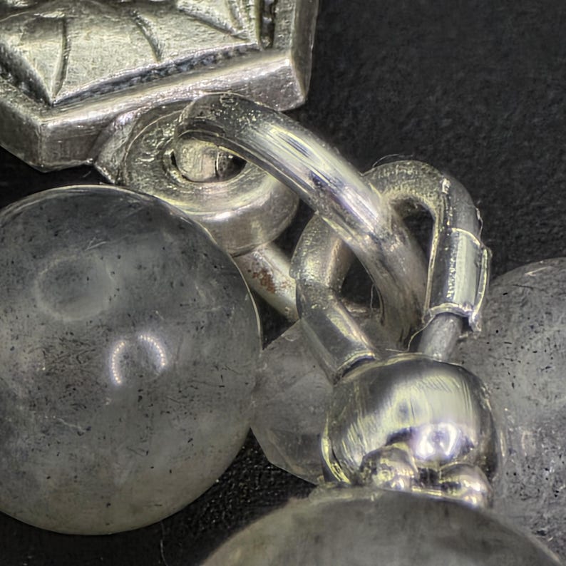 May include: Close-up of a silver bracelet with a decorative clasp and chain links. The bracelet features multiple round, textured beads. The metal has a polished finish, reflecting light against a dark background.