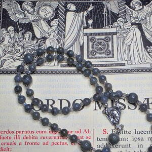 May include: A grey gemstone bead rosary with a silver crucifix pendant. The rosary is laid on a page with religious illustrations and Latin text. The background features detailed black and white drawings of angels and religious figures.