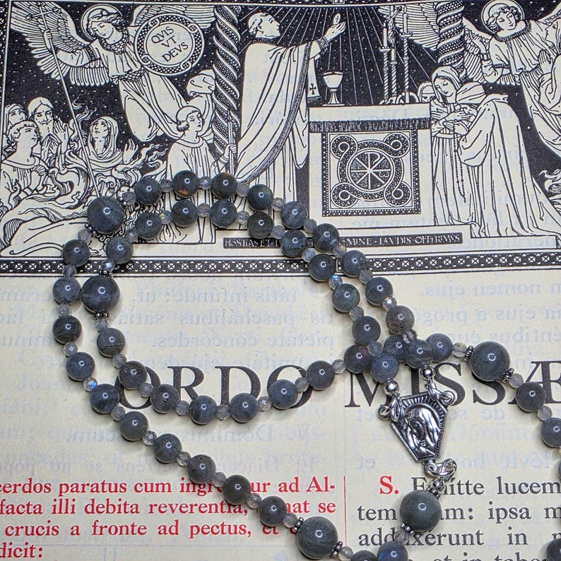 May include: A grey gemstone bead rosary with a silver crucifix pendant. The rosary is laid on a page with religious illustrations and Latin text. The background features detailed black and white drawings of angels and religious figures.