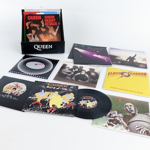 May include: A collection of Queen vinyl records and album covers. The albums include "Sheer Heart Attack", "A Kind of Magic", and "Flash Gordon". A black record holder with the band's name is also present.