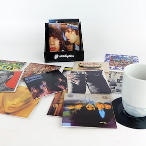 May include: A selection of The Rolling Stones album art coasters. The coasters showcase album covers such as "Black and Blue" and "Aftermath". A black display box with the Rolling Stones logo is also visible.