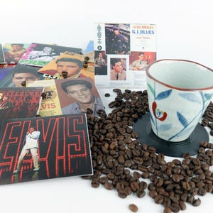 May include: A collection of Elvis Presley memorabilia, including album covers and cards, scattered with coffee beans. A ceramic cup with a floral design sits on a black coaster. The album covers feature images of Elvis in various poses.